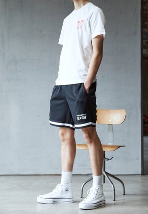 Person, die mit den Händen in den Taschen steht und weiße Converse High-Top-Sneaker, schwarze Converse-Shorts und ein weißes T-Shirt trägt, neben einem Holzstuhl.