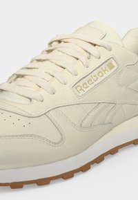 Κοντινή λήψη ενός κρεμ Reebok αθλητικού παπουτσιού με υφή δέρματος, λευκά κορδόνια και καφέ λαστιχένια σόλα.