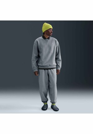 Mężczyzna ubrany w szarą bluzę i spodnie dresowe, czarną koszulę, neonowożółtą czapkę i skarpetki oraz czarne wsuwane buty, stojący bokiem.