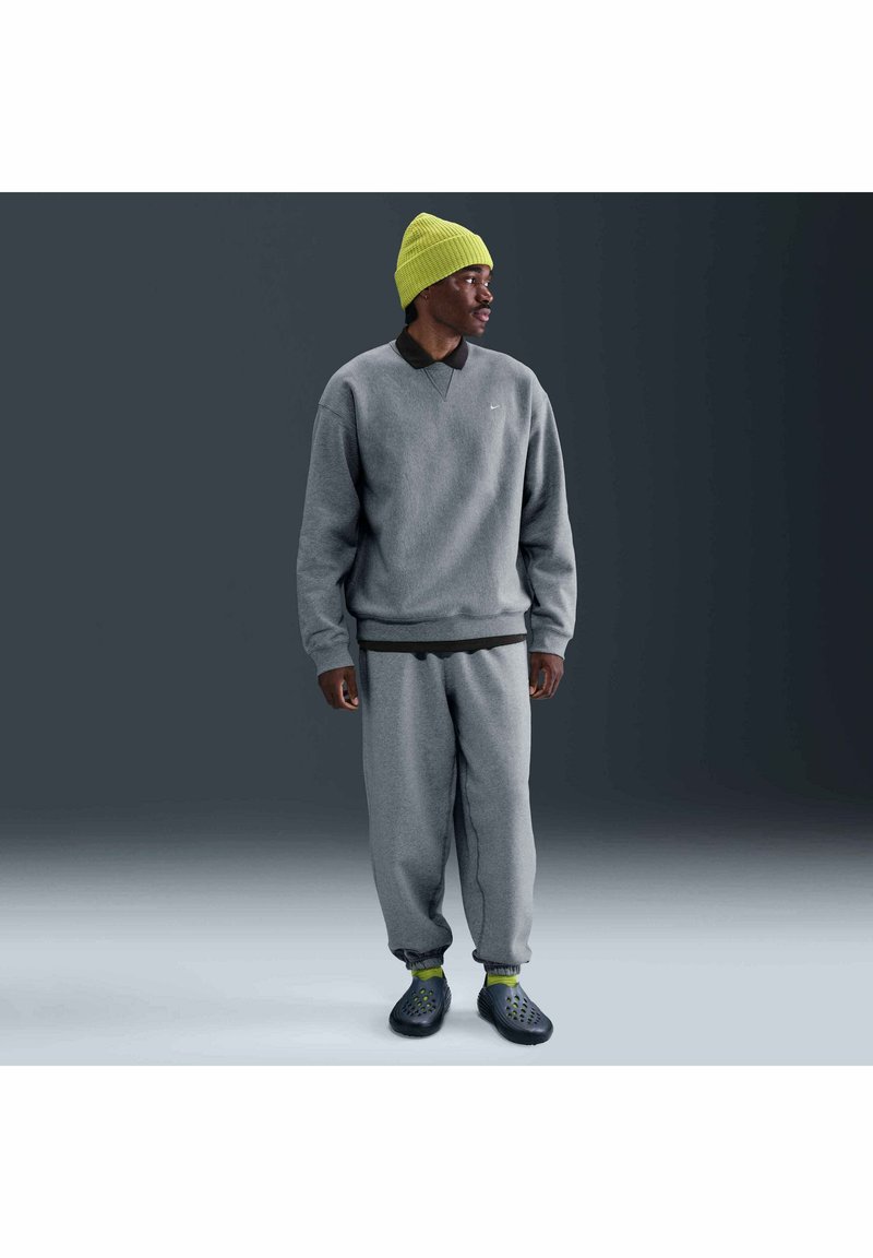 Uomo che indossa una felpa grigia e pantaloni della tuta, una maglietta nera, un berretto e calzini giallo neon, e scarpe slip-on nere, in piedi di lato.