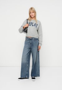 Šedá mikina s logom "Replay", široké modré džínsy a čierne tenisky. Model drží malú čiernu kabelku. Jednoduché pozadie.