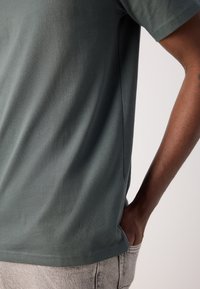 Homme portant une chemise verte à manches courtes avec une main dans la poche d'un jean clair, vu de mi-dos jusqu'au coude.