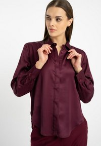 Bordowa bluzka z jedwabistego materiału z długimi rękawami, zapięciem na guziki z przodu i zaokrąglonym dołem. Posiada guzikowe mankiety oraz usztywniony kołnierz.