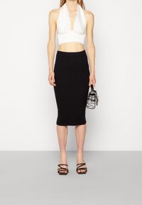 Weißes Satin-Halter-Crop-Top kombiniert mit einem schwarzen gerippten knielangen Rock. Das Model hält eine kleine Handtasche mit Zebra-Print. Schwarze Riemchensandalen runden das Outfit ab.