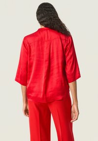 Blusa roja con mangas tres cuartos, hecha de tela suave. Cuenta con un yugo trasero y un dobladillo ligeramente curvado. Combinada con pantalones rojos.