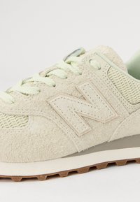 Baskets beige en daim et mesh avec lacets crème, grand logo "N" cousu et détail de semelle en caoutchouc marron sur fond blanc.