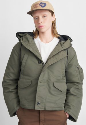 Giacca imbottita verde oliva con cappuccio, dotata di due tasche frontali, chiusura con zip e cordini. Indossata sopra una camicia bianca.