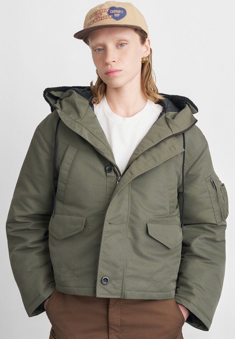 Olīvu zaļa spilventiņu jaka ar kapuci, ar divām priekšējām kabatām, rāvējslēdzēja aizdari un aukliņām. Vilkta virs baltas kreklas.