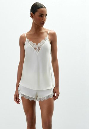 Kvinde iført hvid satin camisole og shorts med blondekant, stående mod en ensfarvet lys baggrund, kiggende til siden.