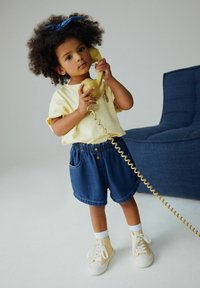 Sárga rövid ujjú póló, rugalmas derékpántos farmer rövidnadrág, fehér zokni és sárga tornacipő. Gyerek vintage telefont tart a kezében.