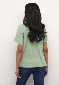 Tricou verde deschis din bumbac, cu mâneci scurte, croi lejer și tiv rotunjit; asortat cu blugi negri din denim. Vedere din spate.