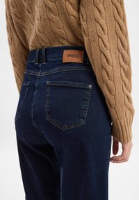 Jeans di denim blu scuro con vita alta, caratterizzati da un'etichetta in pelle con la scritta "ANGELS" e due tasche posteriori con cuciture gialle.