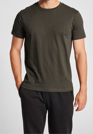 Homme portant un t-shirt uni vert foncé à manches courtes et un pantalon noir, debout, une main dans la poche, devant un fond clair.