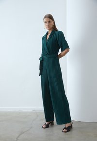 Teal plissé jumpsuit met een wikkelontwerp, korte mouwen, een ceintuur in de taille en een brede pijpen. Gecombineerd met zwarte hoge sandalen.