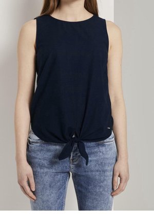 Vrouw draagt een mouwloze navy top met voorstrikdetail en lichtblauwe verweerde spijkerbroek, staand tegen een effen achtergrond.