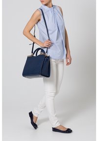 Modrá halenka bez rukávů, bílé úzké džíny a tmavě modrá kabelka s tkanou texturou a zlatými kovovými doplňky, doplněné tmavě modrými balerínami.