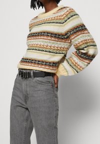 Jersey de cuello redondo con rayas multicolores en verde, marrón, amarillo y crema. Presenta un ajuste holgado y un patrón de punto texturizado. Combinado con jeans grises.
