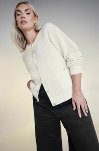 Krämbelagd stickad cardigan med en strukturerad mönster, rundad halsringning, ribbade ärmslut och knäppning. Bärs över mörka byxor.