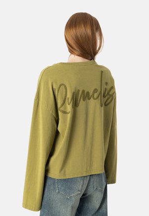Ung person med skulderlangt, rødt hår iført olivengrøn langærmet top med teksten "R. Marcelis" på bagsiden og blå jeans.