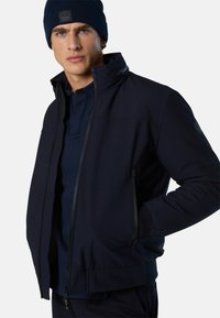 Veste de performance bleu marine avec col montant, fermeture éclair, poches latérales et tissu texturé. Associée à un bonnet en tricot assorti.