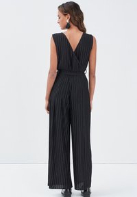 Combinaison noire plissée avec un dos en V profond, des jambes larges et une ceinture à nouer à la taille. Texture lisse avec un design fluide.