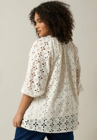 Hvid bluse med broderi og udskårne blomster mønstre; 3/4 ærmer; samlet halsudskæring; løs pasform; båret over blå jeans.