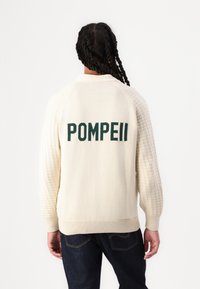 Cream sweater with a textured knit design, featuring green "POMPEII" lettering on the back. Ribbed cuffs and hem. Worn with dark jeans.