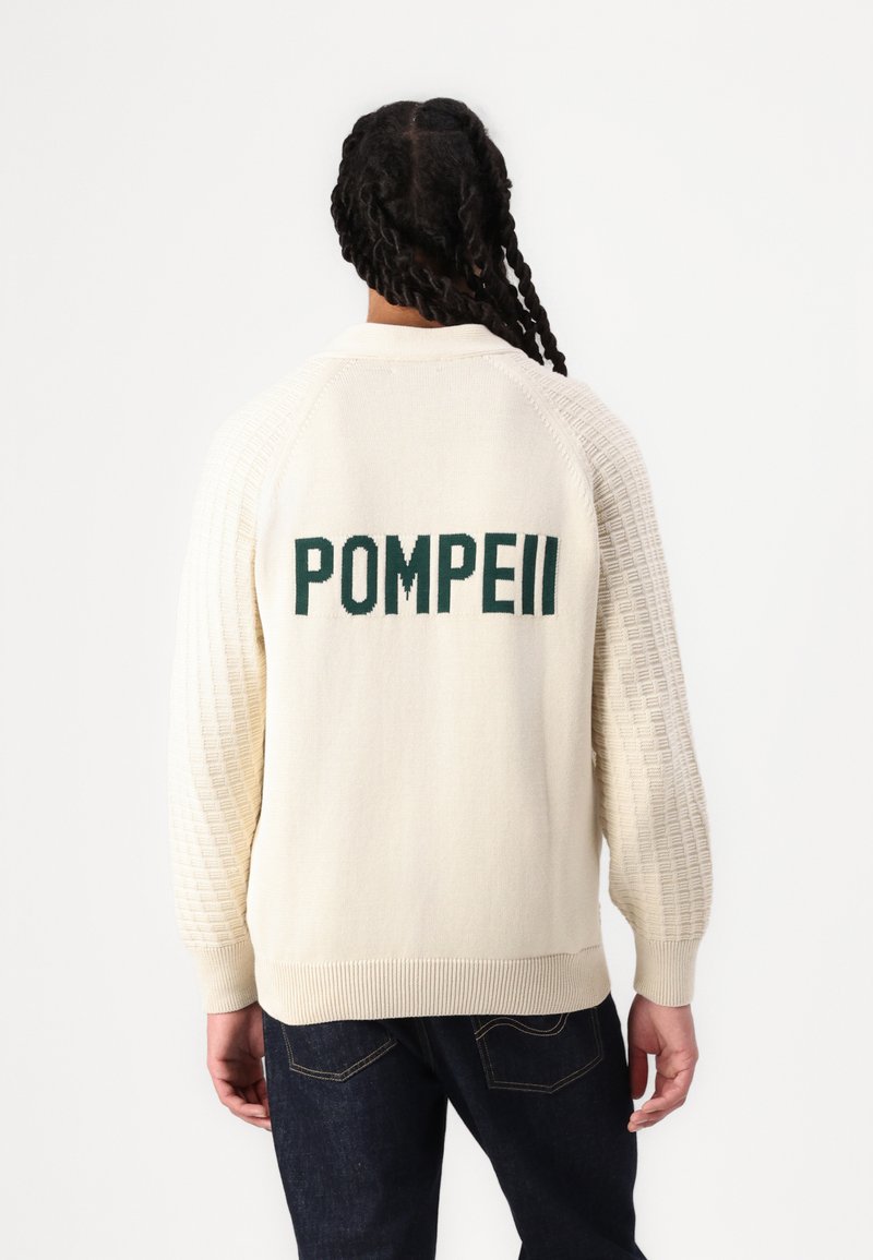 Cream sweater with a textured knit design, featuring green "POMPEII" lettering on the back. Ribbed cuffs and hem. Worn with dark jeans.