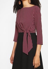 Blouse rouge à motifs avec des manches 3/4, des motifs en diamond, une taille nouée et une jupe noire unie. Tissu texturé avec une finition lisse.