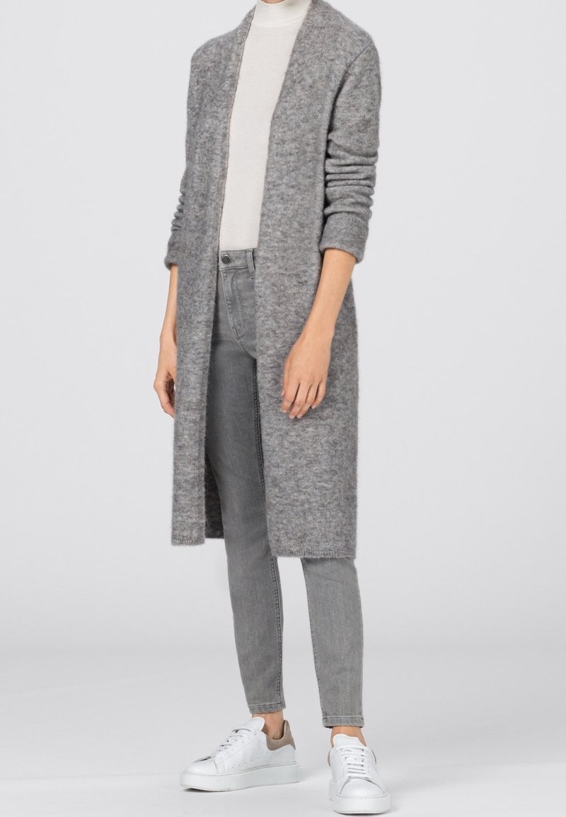 Person wearing a long gray fuzzy cardigan, light gray turtleneck, gray jeans, and white sneakers with beige accents, standing on a plain background.