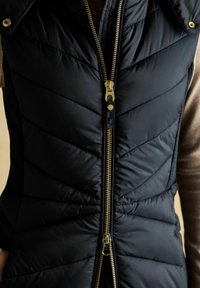 Melnās quiltētas vestes ar divkāršu zelta rāvējslēdzēju, daļēji atvērtas, virs netīras krāsas garām piedurknēm kreklam, redzama vestes zvīņu šuvju raksts.