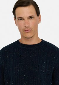 Granatowy, dzianinowy sweter z teksturowanym wzorem i wielokolorowymi flekami. Żebrowany dekolt i mankiety podkreślają design.