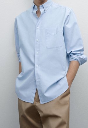 Mand iført lyseblå skjorte med opkrogede ærmer og beige bukser, stående med hænderne i lommerne mod en ensfarvet baggrund.