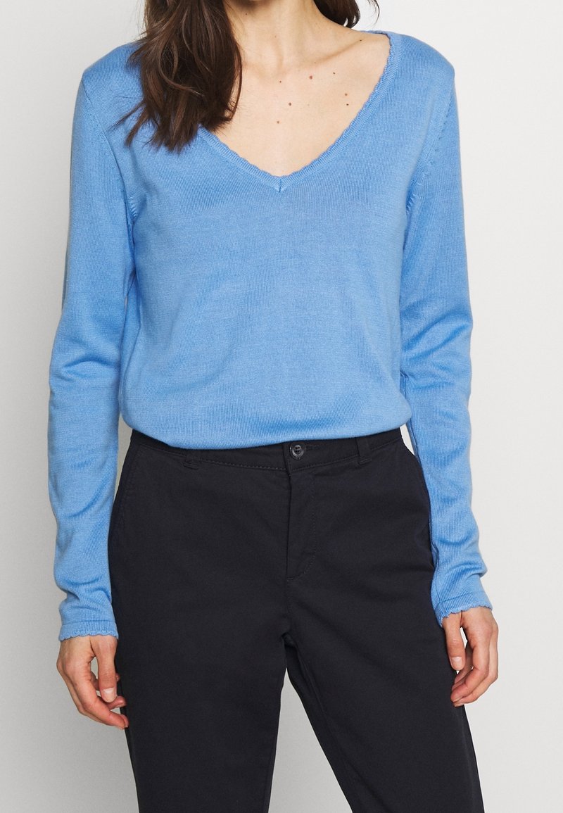 Hellblaues langärmliges Sweater mit V-Ausschnitt, gestrickter Textur und ausgefranstem Saum, kombiniert mit schwarzen Hosen. Eng anliegend.