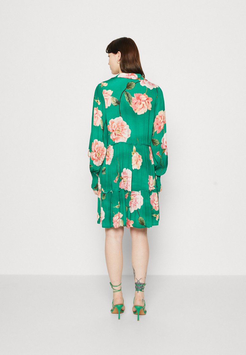 Grünes Blumenkleid mit einem gestuften Design; es hat große rosa Rosen und Blätter, lange Ärmel und eine taillierte Taille. Akzentuiert mit schnürenden grünen High Heels.