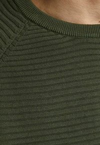 Pull vert en maille côtelée avec une surface texturée et un col rond, mettant en avant un gros plan des détails complexes du tissu.