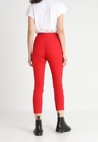 Rode high-waisted broek met een strakke pasvorm, cropped lengte en een gladde textuur, gecombineerd met zwarte enkellaarsjes. Witte top met korte mouwen zichtbaar.