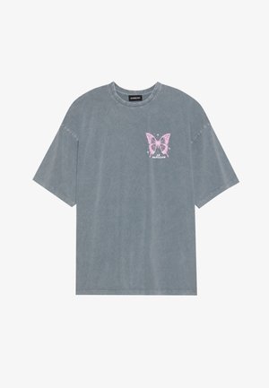 T-shirt gris surdimensionné en coton doux avec des manches courtes. Présente un graphique de papillon rose sur la partie gauche de la poitrine.