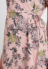 Gros plan sur une robe portefeuille rose avec une taille nouée et un motif floral arborant des fleurs jaunes, blanches et roses avec des feuilles vertes.