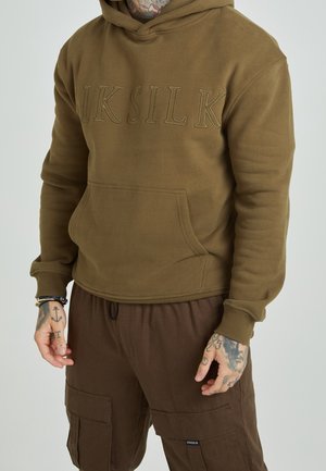 Mand iført brun hættetrøje med "SIKSILK" broderi og brune cargobukser, synlige håndtattooer og armbånd, neutral baggrund.