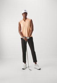 Polo in light orange with a collar, paired with black trousers. White sport shoes and a white cap. Holding a golf club. Minimalist background.