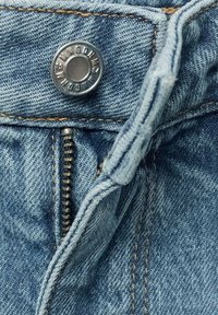 Tessuto denim azzurro chiaro con cuciture gialle a contrasto, dotato di un bottone in metallo argentato e zip in vita. Dettaglio ravvicinato.
