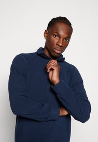 Pullover in pile blu navy con colletto a zip, maniche lunghe e texture liscia. Presenta un piccolo logo sul petto e dettagli di cucitura minimali.