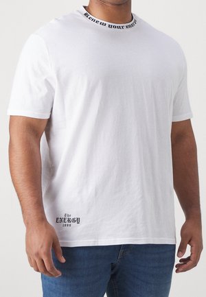Homme portant un T-shirt blanc à manches courtes avec le texte noir « Renouvez votre énergie » sur le col et « The Energy 1999 » près de l'ourlet, associé à un jean bleu.