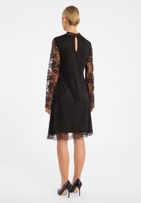 Zwarte kanten jurk met lange doorschijnende mouwen, sleutelgatdetail op de rug, geschulpte zoom en een aansluitende pasvorm, gecombineerd met zwarte hoge hakken.