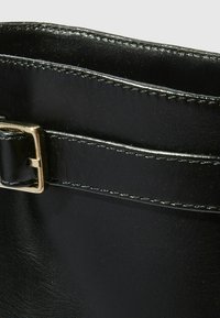 Dettaglio di una borsa in pelle nera con una fibbia in metallo dorato, texture liscia e cuciture bianche a contrasto lungo i bordi.