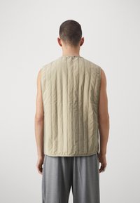 Beżowa bezrękawnikowa kamizelka z pionową pikowaną teksturą, okrągłym dekoltem i luźnym krojem, noszona na jasnoszarych spodniach.