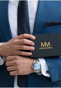 Silberne Armbanduhr mit blauem Zifferblatt und schwarzem Lünette, getragen an einem elegant gekleideten Handgelenk, das eine schwarze Box mit goldener Schrift hält, "MASSI MORINO."
