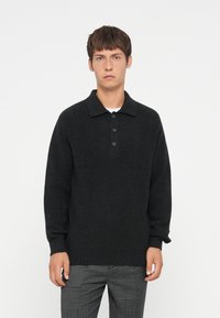 Zwart gebreid pullover met een kraag en sluiting met vier knopen. Lange mouwen en gestructureerde stof. Gepaarde met grijs geruite broek.
