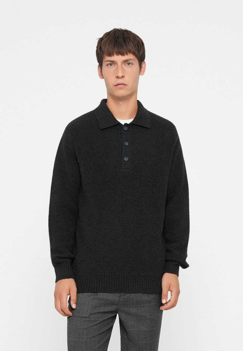 Zwart gebreid pullover met een kraag en sluiting met vier knopen. Lange mouwen en gestructureerde stof. Gepaarde met grijs geruite broek.
